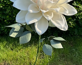 Pode incluir: Uma grande flor de dália artificial branca com um centro rosa, em um caule verde com folhas, em pé sobre uma base verde em uma área gramada.