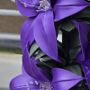 Puede incluir: Un arreglo vertical de flores de lirio artificiales de color morado con p&eacute;talos lisos y mate. Cada flor presenta estambres oscuros y delgados con peque&ntilde;as cuentas transparentes y reflectantes. Hojas texturizadas de color verde oscuro se intercalan entre las flores.