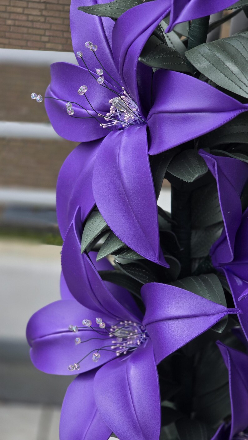 Puede incluir: Un arreglo vertical de flores de lirio artificiales de color morado con p&eacute;talos lisos y mate. Cada flor presenta estambres oscuros y delgados con peque&ntilde;as cuentas transparentes y reflectantes. Hojas texturizadas de color verde oscuro se intercalan entre las flores.