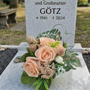 Puede incluir: Una lápida de granito gris claro con el texto "Hier ruht unsere geliebte Mutter und Großmutter GÖTZ * 1941 † 2024" que presenta un arreglo floral de rosas color melocotón, hojas verdes y una pequeña figura decorativa de ángel.