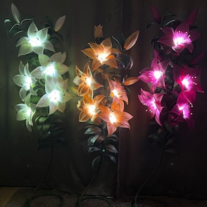 Puede incluir: Tres l&aacute;mparas de pie altas con forma de lirio iluminadas. La l&aacute;mpara de la izquierda brilla con luz blanca, la central con luz naranja c&aacute;lida y la de la derecha con luz rosa vibrante. Cada l&aacute;mpara presenta tallos y hojas verdes.