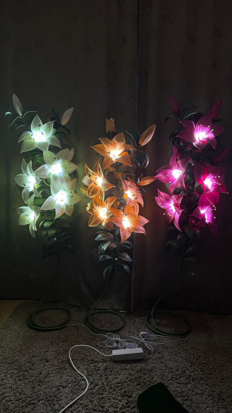 Puede incluir: Tres l&aacute;mparas de pie altas con forma de lirio iluminadas. La l&aacute;mpara de la izquierda brilla con luz blanca, la central con luz naranja c&aacute;lida y la de la derecha con luz rosa vibrante. Cada l&aacute;mpara presenta tallos y hojas verdes.