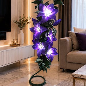 Puede incluir: Una l&aacute;mpara de pie alta con forma de racimo de flores de lirio de color p&uacute;rpura vibrante con hojas verdes. Los p&eacute;talos est&aacute;n iluminados desde el interior, emitiendo un brillo blanco intenso. La base es un tallo verde enrollado con cable.