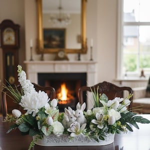 Könnte beinhalten: Weißes Blumengesteck mit zwei Hasenfiguren und dekorativen Eiern in einem weißen, spitzenbesetzten Behälter. Die Anordnung umfasst weiße Blumen und Grün, die auf einem dunklen Holztisch platziert sind. Ein Kamin und ein Spiegel befinden sich im Hintergrund.