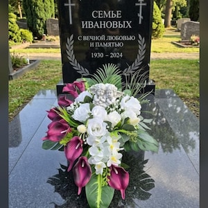 May include: A black granite headstone with engraved text reading "СЕМЬЕ ИВАНОВЫХ", "С ВЕЧНОЙ ЛЮБОВЬЮ И ПАМЯТЬЮ", and "1930 – 2024", flanked by two crosses. A floral arrangement of white roses, purple calla lilies, and a small white angel figurine sits in front.