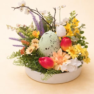 Könnte beinhalten: Osterarrangement in einer weißen Keramikschale. Es zeigt ein gesprenkeltes grünes Ei, rote und weiße Tulpen, gelbe Gänseblümchen und verschiedene andere bunte Blumen und Grün. Das Arrangement befindet sich vor einem hellgelben Hintergrund.