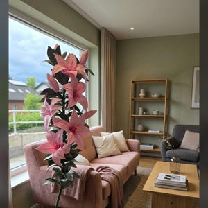 Puede incluir: Una planta artificial alta de pie que presenta m&uacute;ltiples flores de lirio de color rosa vibrante con hojas de color verde oscuro sobre un tallo delgado de color verde oscuro que se curva en una base circular con un cable el&eacute;ctrico blanco.