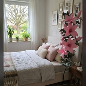 Puede incluir: Una l&aacute;mpara de pie alta con forma de gran flor de lirio rosa con hojas verdes y un tallo oscuro. La l&aacute;mpara presenta varios p&eacute;talos de color rosa en flor y est&aacute; colocada junto a una cama con almohadas claras y una manta gris.