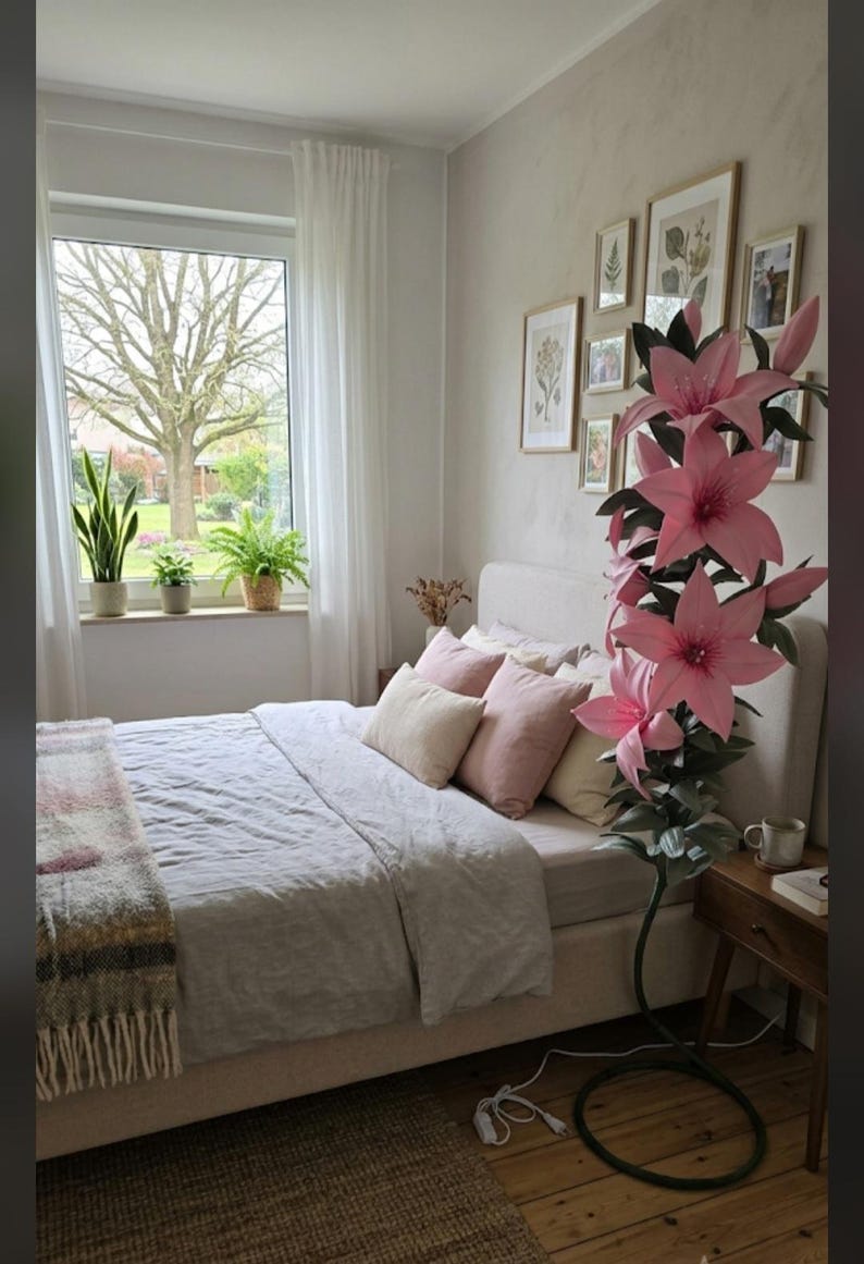 Puede incluir: Una l&aacute;mpara de pie alta con forma de gran flor de lirio rosa con hojas verdes y un tallo oscuro. La l&aacute;mpara presenta varios p&eacute;talos de color rosa en flor y est&aacute; colocada junto a una cama con almohadas claras y una manta gris.