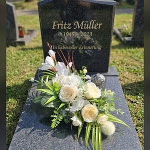 May include: A polished black granite headstone with the gold-engraved text Fritz Müller, 1945-2023, and In liebevoller Erinnerung. A floral arrangement of white roses, lilies, and green foliage rests on the base of the grave marker.