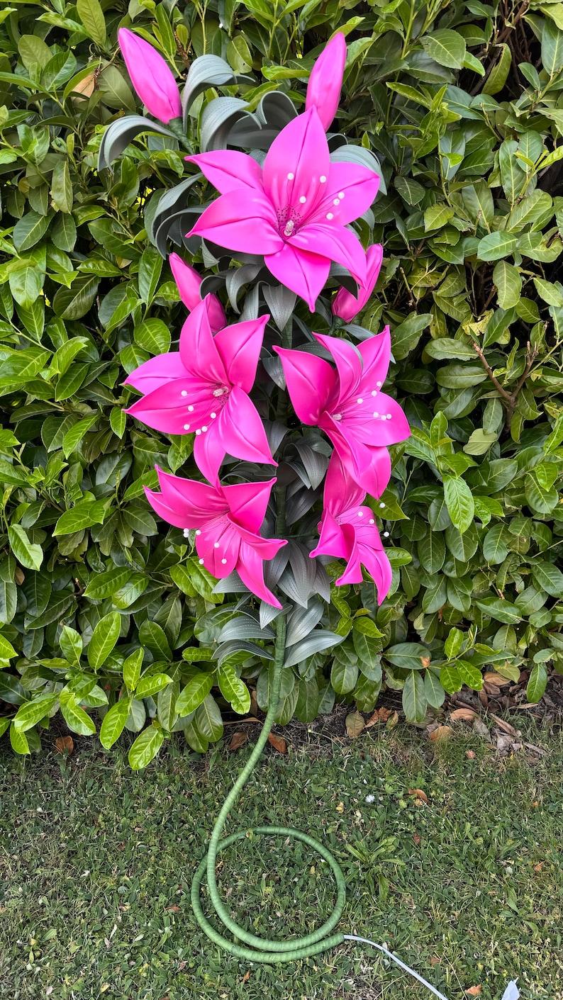 Puede incluir: Una planta artificial decorativa alta que presenta m&uacute;ltiples flores de lirio de color rosa vibrante con estambres de punta blanca y hojas de color verde oscuro. El tallo est&aacute; envuelto en material verde y se enrosca en una base circular con un cable blanco.