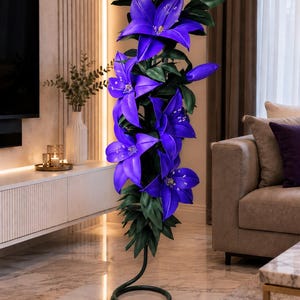 Puede incluir: Una l&aacute;mpara de pie alta con forma de racimo de flores de lirio de color p&uacute;rpura vibrante con hojas de color verde oscuro. La l&aacute;mpara cuenta con una base negra en espiral que descansa en el suelo, con un cable blanco fino desde la base.