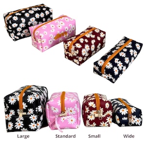 Puede incluir: Una colección de estuches con estampado de margaritas en varios tamaños y colores. Los estuches presentan un estampado de margaritas blancas sobre fondos negros, rosas, burdeos y azul marino. Cada estuche tiene una cremallera naranja.