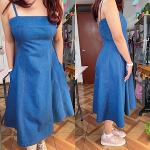 May include: A blue denim dress with a square neckline and spaghetti straps. The dress has a fitted bodice, a flared skirt, and pockets. The seams are highlighted with a contrasting orange thread. The dress is knee-length.