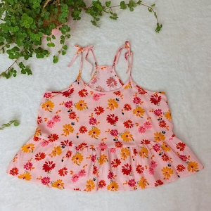 Puede incluir: Una blusa rosa sin mangas con un dobladillo con volantes y un estampado floral. La blusa tiene tirantes finos que se atan en los hombros.