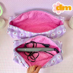 Puede incluir: Dos neceseres con cremallera abiertos. El exterior es un estampado de cuadros morados claros con pequeñas ilustraciones de fresas rojas. El interior es de tela rosa acolchada. Una bolsa contiene una plancha de pelo Remington gris y un cable negro.