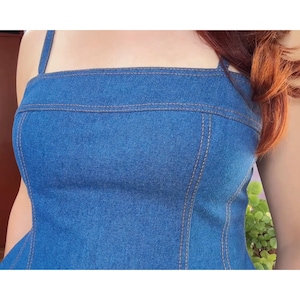May include: A blue denim top with a square neckline and thin straps. The top features visible tan stitching along the seams and a fitted bodice. The fabric has a classic denim texture.