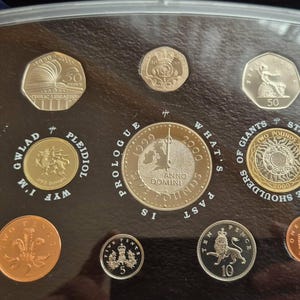 Puede incluir: Colección de monedas británicas de varios valores, incluyendo una moneda de 50 peniques y una moneda de 2 libras, expuestas en una funda protectora. Las monedas presentan diseños e inscripciones detalladas.