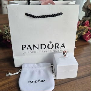 K&ouml;nnte beinhalten: Eine wei&szlig;e Geschenkt&uuml;te mit schwarzen Seilgriffen und dem Logo "PANDORA UNFORGETTABLE MOMENTS". Eine wei&szlig;e Schachtel und ein kleiner wei&szlig;er Beutel mit dem Logo "PANDORA" befinden sich neben der T&uuml;te. Ein Ring mit roten und silbernen Akzenten liegt auf der Schachtel.