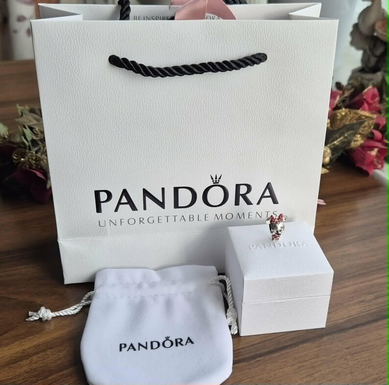 K&ouml;nnte beinhalten: Eine wei&szlig;e Geschenkt&uuml;te mit schwarzen Seilgriffen und dem Logo "PANDORA UNFORGETTABLE MOMENTS". Eine wei&szlig;e Schachtel und ein kleiner wei&szlig;er Beutel mit dem Logo "PANDORA" befinden sich neben der T&uuml;te. Ein Ring mit roten und silbernen Akzenten liegt auf der Schachtel.