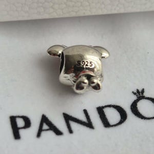 K&ouml;nnte beinhalten: Ein silberner Charm in Form eines B&auml;ren mit kleinen Ohren und F&uuml;&szlig;en. Der Charm ist mit "925" gestempelt und liegt auf einer wei&szlig;en Oberfl&auml;che mit dem schwarzen Aufdruck "PANDORA".