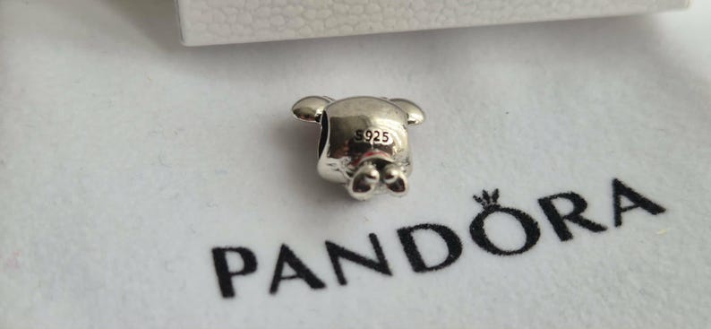 K&ouml;nnte beinhalten: Ein silberner Charm in Form eines B&auml;ren mit kleinen Ohren und F&uuml;&szlig;en. Der Charm ist mit "925" gestempelt und liegt auf einer wei&szlig;en Oberfl&auml;che mit dem schwarzen Aufdruck "PANDORA".