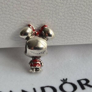 K&ouml;nnte beinhalten: Ein silberner Charm-Anh&auml;nger in Form einer Zeichentrickfigur mit roter Schleife und Kleid. Der Anh&auml;nger befindet sich vor einem wei&szlig;en Hintergrund und einer wei&szlig;en Box.