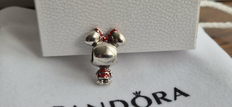 K&ouml;nnte beinhalten: Ein silberner Charm-Anh&auml;nger in Form einer Zeichentrickfigur mit roter Schleife und Kleid. Der Anh&auml;nger befindet sich vor einem wei&szlig;en Hintergrund und einer wei&szlig;en Box.