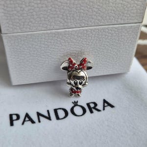 K&ouml;nnte beinhalten: Ein silberner Minnie-Maus-Charm mit roter Schleife und Kleid, auf einem wei&szlig;en Beutel mit dem "PANDORA"-Logo. Der Charm befindet sich neben einer wei&szlig;en, strukturierten Box.