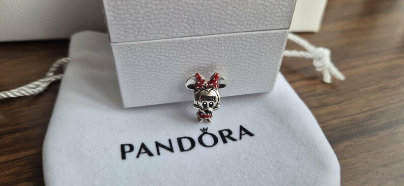 K&ouml;nnte beinhalten: Ein silberner Minnie-Maus-Charm mit roter Schleife und Kleid, auf einem wei&szlig;en Beutel mit dem "PANDORA"-Logo. Der Charm befindet sich neben einer wei&szlig;en, strukturierten Box.