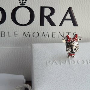 K&ouml;nnte beinhalten: Ein silberner Minnie Mouse-Charm mit roter Schleife und Kleid, pr&auml;sentiert auf einer wei&szlig;en Pandora-Box. Der Charm ist mit schwarzen und roten Akzenten detailliert. Im Hintergrund eine wei&szlig;e Box mit dem Pandora-Logo.