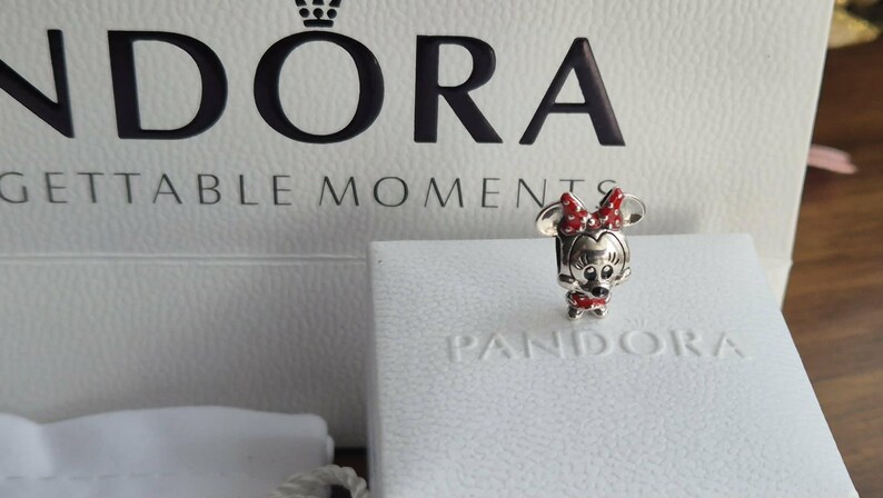 K&ouml;nnte beinhalten: Ein silberner Minnie Mouse-Charm mit roter Schleife und Kleid, pr&auml;sentiert auf einer wei&szlig;en Pandora-Box. Der Charm ist mit schwarzen und roten Akzenten detailliert. Im Hintergrund eine wei&szlig;e Box mit dem Pandora-Logo.