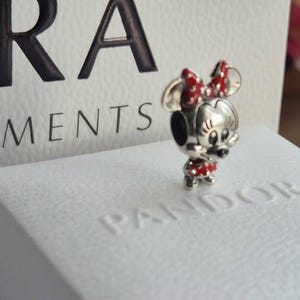 K&ouml;nnte beinhalten: Ein silberner Minnie Mouse Charm mit roter und wei&szlig;er Polka-Dot-Schleife und Kleid. Der Charm steht auf einer wei&szlig;en Box mit dem gepr&auml;gten Wort "Pandora". Der Hintergrund enth&auml;lt das Wort "Moments".
