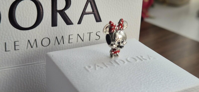 K&ouml;nnte beinhalten: Ein silberner Minnie Mouse Charm mit roter und wei&szlig;er Polka-Dot-Schleife und Kleid. Der Charm steht auf einer wei&szlig;en Box mit dem gepr&auml;gten Wort "Pandora". Der Hintergrund enth&auml;lt das Wort "Moments".