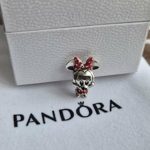 K&ouml;nnte beinhalten: Ein silberner Minnie-Maus-Charm mit roter Schleife und Kleid, auf einem wei&szlig;en Beutel mit dem Logo "PANDORA". Der Charm ist detailliert und vor einer wei&szlig;en Box.