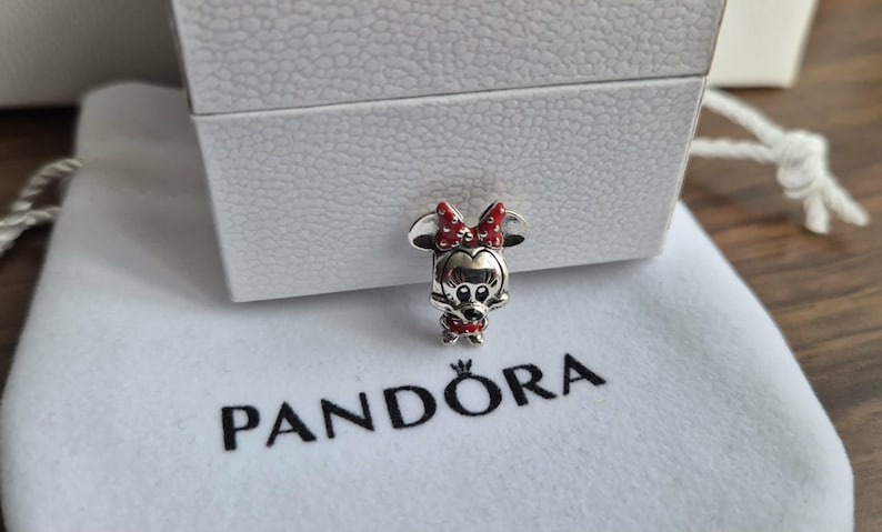 K&ouml;nnte beinhalten: Ein silberner Minnie-Maus-Charm mit roter Schleife und Kleid, auf einem wei&szlig;en Beutel mit dem Logo "PANDORA". Der Charm ist detailliert und vor einer wei&szlig;en Box.