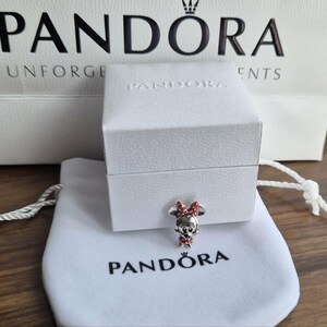 K&ouml;nnte beinhalten: Ein Pandora-Charm mit Minnie Mouse mit roter Schleife und Kleid, pr&auml;sentiert auf einem wei&szlig;en Beutel und einer Box. Das Pandora-Logo ist auf dem Beutel und der Einkaufstasche im Hintergrund zu sehen.