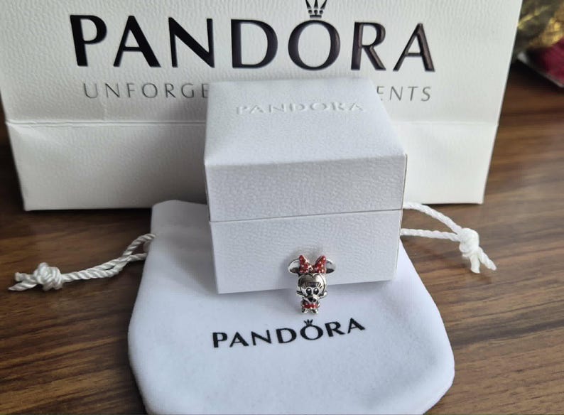 K&ouml;nnte beinhalten: Ein Pandora-Charm mit Minnie Mouse mit roter Schleife und Kleid, pr&auml;sentiert auf einem wei&szlig;en Beutel und einer Box. Das Pandora-Logo ist auf dem Beutel und der Einkaufstasche im Hintergrund zu sehen.