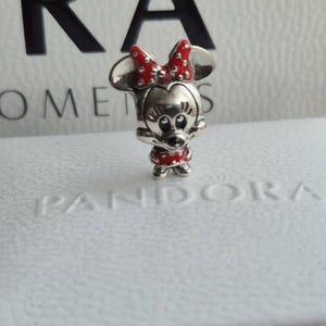 K&ouml;nnte beinhalten: Ein silberner Minnie-Mouse-Charm mit roter Schleife und Kleid. Der Charm steht auf einer wei&szlig;en Schachtel mit dem Aufdruck "PANDORA". Der Hintergrund umfasst eine wei&szlig;e Schachtel mit den Aufdrucken "ORA" und "LE MOMENTS".
