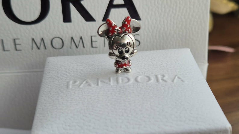 K&ouml;nnte beinhalten: Ein silberner Minnie-Mouse-Charm mit roter Schleife und Kleid. Der Charm steht auf einer wei&szlig;en Schachtel mit dem Aufdruck "PANDORA". Der Hintergrund umfasst eine wei&szlig;e Schachtel mit den Aufdrucken "ORA" und "LE MOMENTS".