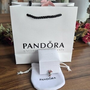 K&ouml;nnte beinhalten: Eine wei&szlig;e Einkaufstasche mit schwarzen Seilgriffen und dem Markennamen "PANDORA" in Schwarz aufgedruckt. Eine kleine wei&szlig;e Schachtel und ein wei&szlig;er Kordelzugbeutel mit dem "PANDORA"-Logo befinden sich davor. Ein Charm mit rosa und roten Akzenten ist sichtbar.
