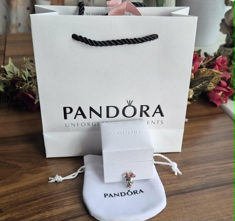 K&ouml;nnte beinhalten: Eine wei&szlig;e Einkaufstasche mit schwarzen Seilgriffen und dem Markennamen "PANDORA" in Schwarz aufgedruckt. Eine kleine wei&szlig;e Schachtel und ein wei&szlig;er Kordelzugbeutel mit dem "PANDORA"-Logo befinden sich davor. Ein Charm mit rosa und roten Akzenten ist sichtbar.