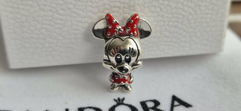 K&ouml;nnte beinhalten: Ein silberner Charm-Anh&auml;nger mit Minnie Mouse, mit roter Schleife und Kleid mit Tupfen. Der Charm hat ein detailliertes Design mit schwarzen Augen und einem l&auml;chelnden Ausdruck. Ein Sammlerst&uuml;ck.