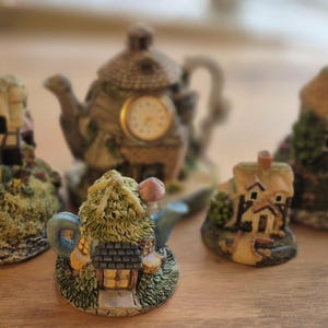 Peut inclure: Une collection de figurines miniatures de cottages et une horloge en forme de théière. Les cottages présentent des détails tels que des toits de chaume, des fenêtres et des portes, entourés de verdure. L'horloge théière a un cadran. La palette de couleurs comprend le beige, le vert et le marron.