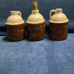 May include: Three brown and white ceramic jugs with cork stoppers. The jugs are decorated with handwritten text that reads "Morgan County Sorghum Festival 1981", "Morgan County Sorghum Festival 1982", and "County Sorghum Festival 1976".
