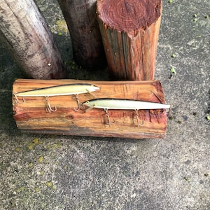 May include: Two gold fishing lures with treble hooks on a wooden surface. The lures are about 6 inches long and have a silver finish.