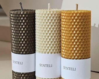 Op de afbeelding: Drie honingraat-kaarsen in verschillende kleuren: donkerbruin, crème en goud. Elke kaars heeft een label met de tekst "SANTELI".