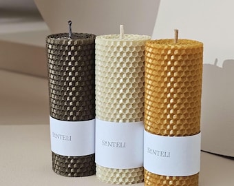 Op de afbeelding: Drie honingraat kaarsen in bruin, crème en goud. De kaarsen zijn gelabeld met "SANTELI" en zijn in een rij op een licht beige oppervlak gerangschikt.