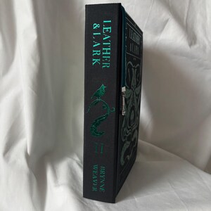 Puede incluir: Un libro de tapa dura negro con el t&iacute;tulo "LEATHER & LARK" de Brynne Weaver. La portada y el lomo presentan letras e ilustraciones verdes, incluyendo un p&aacute;jaro y una serpiente. Se ve una cremallera verde.