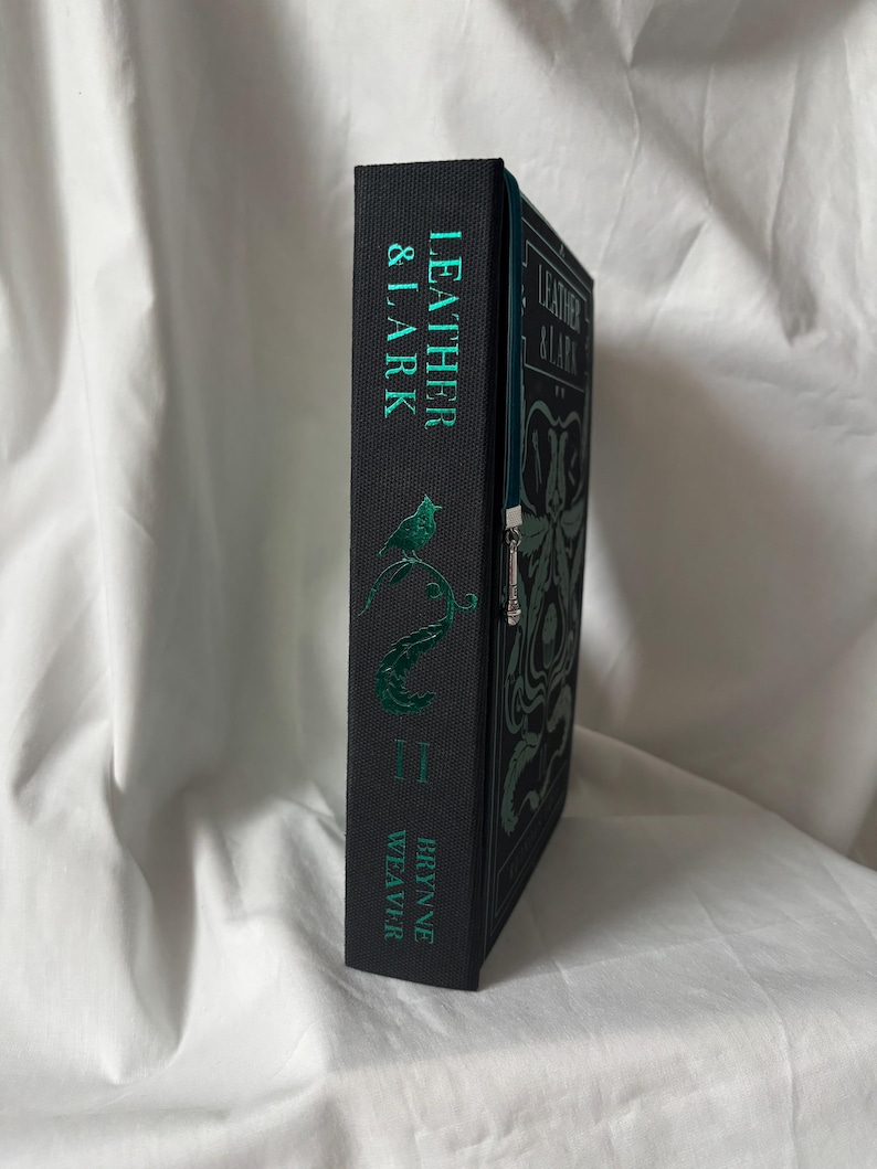 Puede incluir: Un libro de tapa dura negro con el t&iacute;tulo "LEATHER & LARK" de Brynne Weaver. La portada y el lomo presentan letras e ilustraciones verdes, incluyendo un p&aacute;jaro y una serpiente. Se ve una cremallera verde.